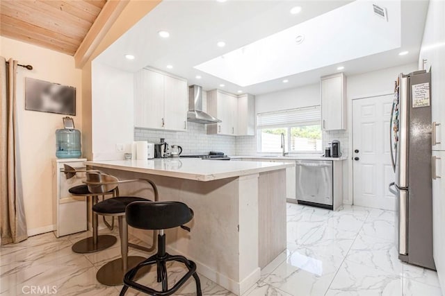 kitchen with wall chimney exhaust hood, stainless steel appliances, backsplash, kitchen peninsula, and a kitchen bar