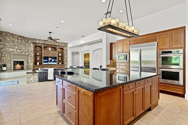 kitchen featuring pendant lighting, a center island, built in appliances, dark stone countertops, and a fireplace