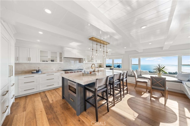 kitchen featuring beam ceiling, a water view, white cabinets, light hardwood / wood-style floors, and an island with sink