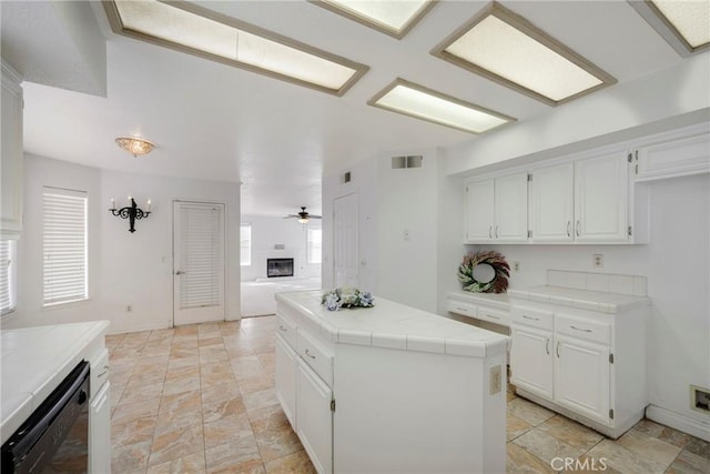 kitchen with dishwasher, a glass covered fireplace, tile countertops, a kitchen island, and white cabinetry