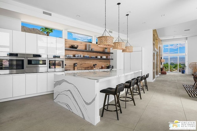 kitchen featuring white cabinets, a center island with sink, hanging light fixtures, and light stone countertops