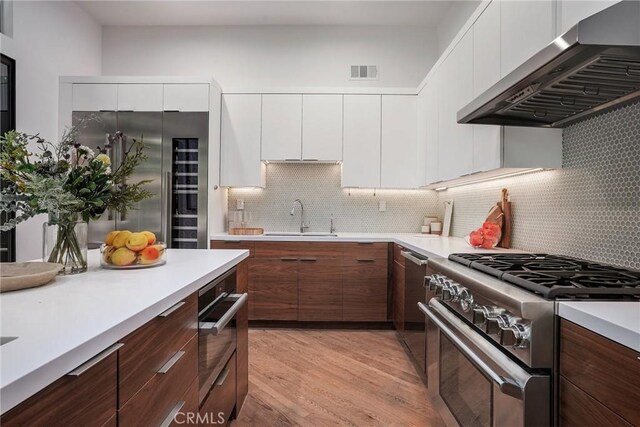 kitchen with high end appliances, sink, light wood-type flooring, white cabinetry, and extractor fan