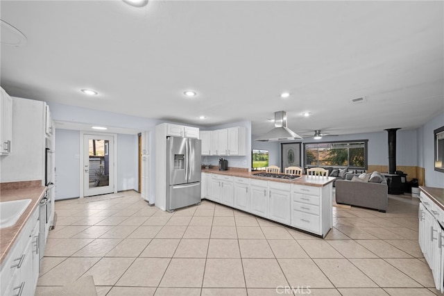 kitchen featuring a wood stove, white cabinets, wall chimney range hood, kitchen peninsula, and stainless steel appliances