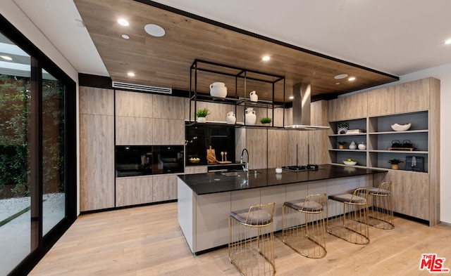 kitchen featuring sink, stainless steel gas cooktop, wall chimney exhaust hood, light hardwood / wood-style flooring, and a kitchen breakfast bar