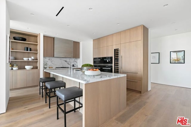 kitchen with an island with sink, tasteful backsplash, light hardwood / wood-style flooring, a breakfast bar area, and light stone countertops