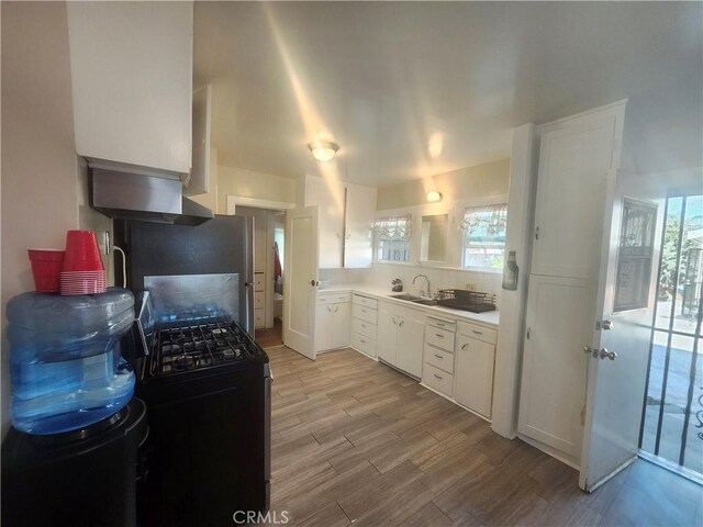 kitchen with black gas range oven, wall chimney range hood, sink, light hardwood / wood-style flooring, and white cabinetry