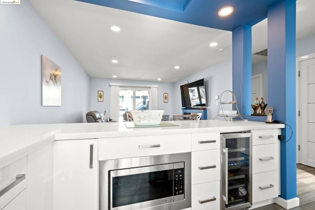 kitchen featuring stainless steel microwave, kitchen peninsula, wine cooler, hardwood / wood-style floors, and white cabinetry