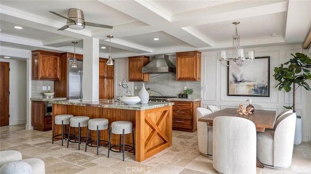 kitchen with beam ceiling, wall chimney exhaust hood, hanging light fixtures, coffered ceiling, and a kitchen island with sink