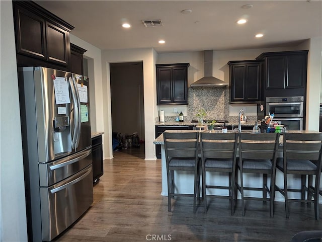 kitchen with a kitchen island with sink, dark wood-type flooring, wall chimney exhaust hood, tasteful backsplash, and stainless steel appliances