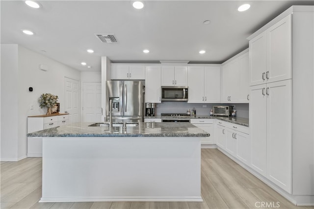 kitchen with an island with sink, dark stone counters, light wood-type flooring, white cabinetry, and appliances with stainless steel finishes