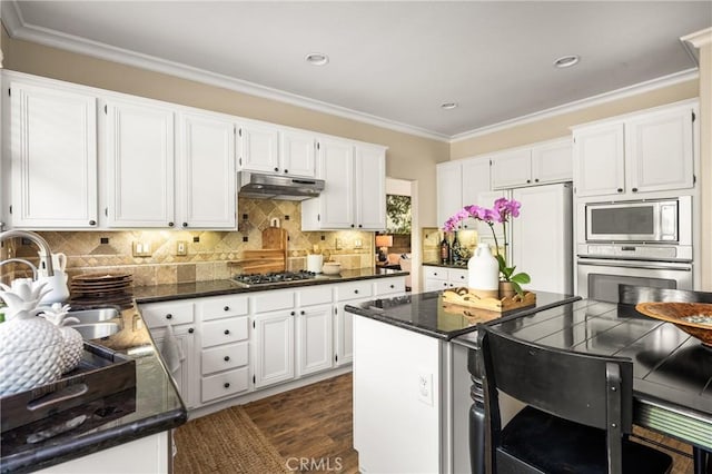 kitchen with white cabinets, built in appliances, under cabinet range hood, crown molding, and backsplash