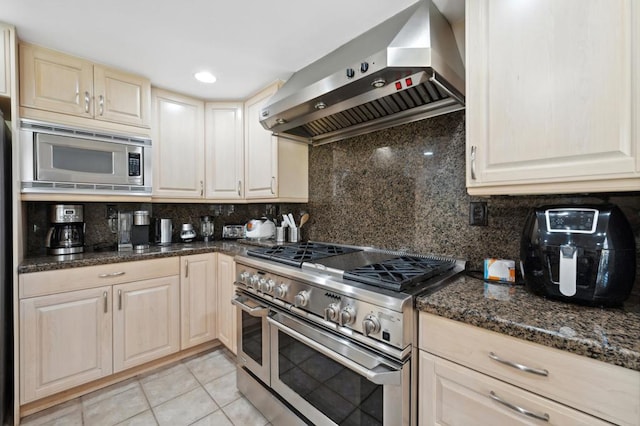 kitchen with decorative backsplash, wall chimney range hood, dark stone countertops, light tile patterned floors, and appliances with stainless steel finishes