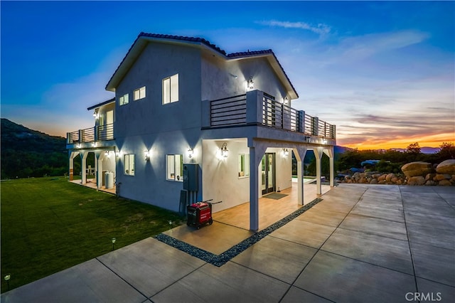 back house at dusk with a patio, a lawn, and a balcony