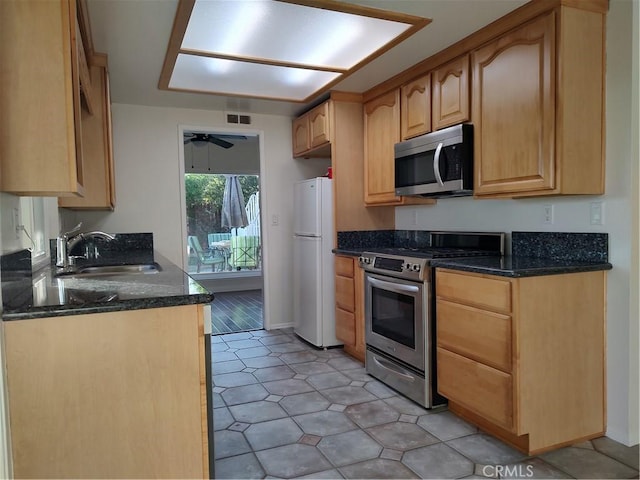 kitchen with dark stone counters, stainless steel appliances, ceiling fan, sink, and light brown cabinets