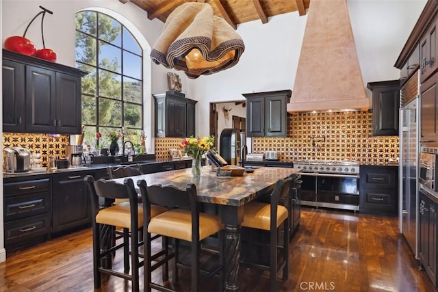 kitchen featuring custom exhaust hood, backsplash, an island with sink, stainless steel range, and beam ceiling