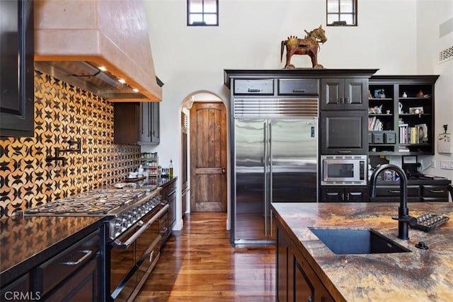 kitchen featuring sink, dark hardwood / wood-style floors, built in appliances, dark stone countertops, and custom exhaust hood