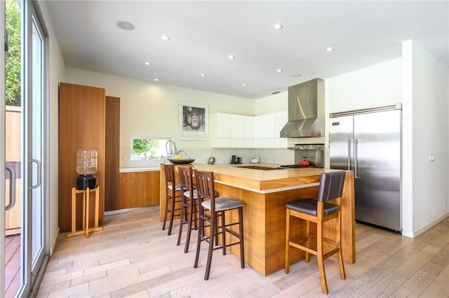 kitchen featuring light wood finished floors, light countertops, backsplash, wall chimney range hood, and built in refrigerator