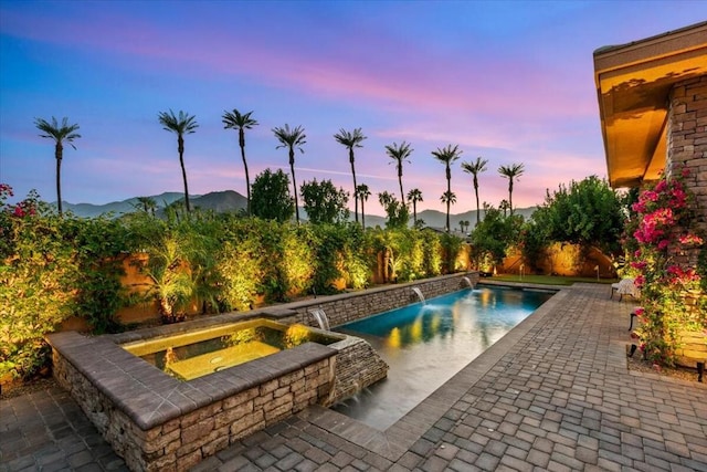 pool at dusk with pool water feature, a mountain view, a patio area, and an in ground hot tub