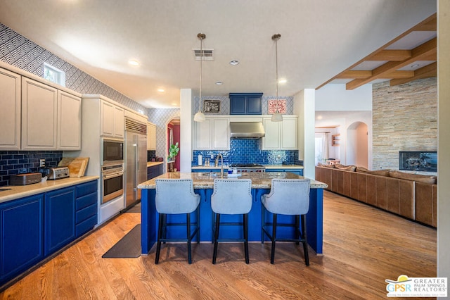 kitchen featuring built in appliances, a center island with sink, hanging light fixtures, and wall chimney exhaust hood