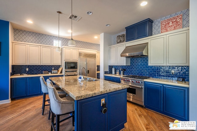 kitchen with wall chimney range hood, blue cabinets, built in appliances, an island with sink, and light wood-type flooring