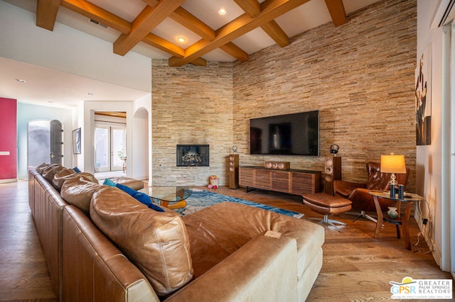 living room featuring beamed ceiling, a towering ceiling, light wood-type flooring, and a stone fireplace