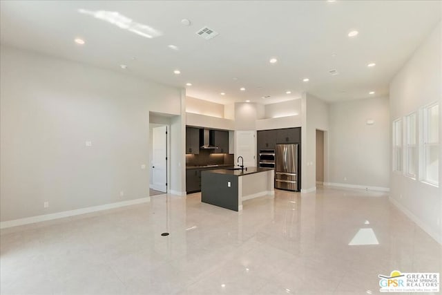 kitchen with a high ceiling, wall chimney exhaust hood, an island with sink, sink, and appliances with stainless steel finishes