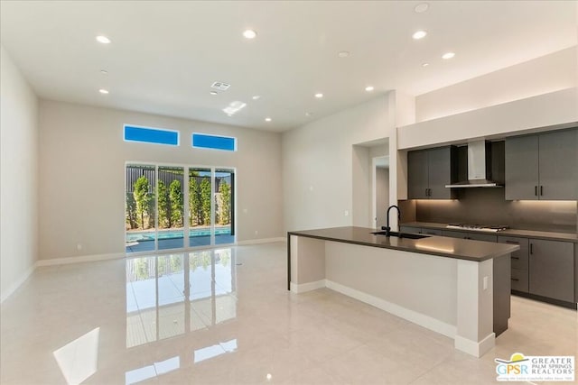 kitchen featuring stainless steel gas cooktop, wall chimney exhaust hood, an island with sink, gray cabinetry, and sink