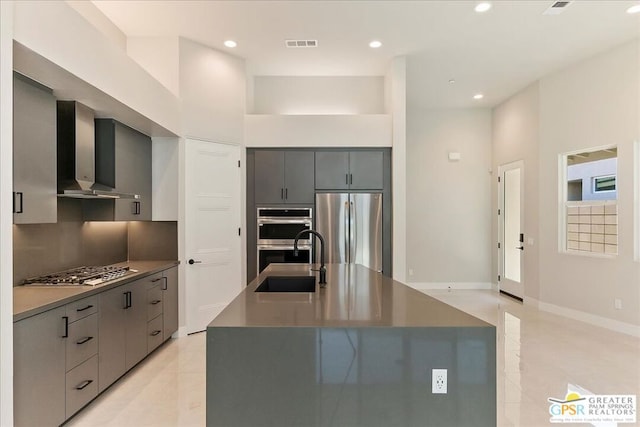 kitchen featuring wall chimney range hood, an island with sink, sink, light tile patterned floors, and appliances with stainless steel finishes