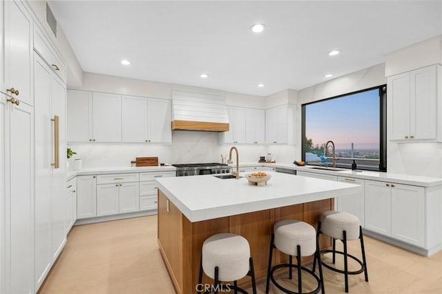 kitchen with custom exhaust hood, a kitchen breakfast bar, stainless steel stove, an island with sink, and white cabinetry