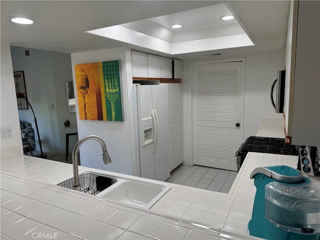 kitchen with tile counters, white fridge with ice dispenser, a tray ceiling, sink, and white cabinetry