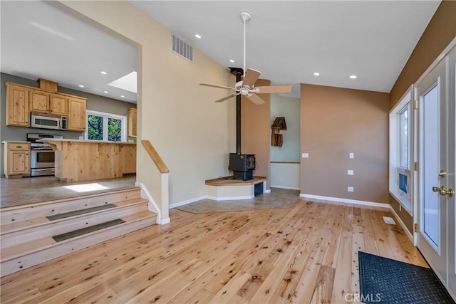 interior space with appliances with stainless steel finishes, light wood-type flooring, ceiling fan, light brown cabinets, and a wood stove