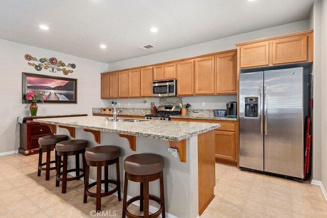 kitchen with stainless steel appliances, an island with sink, sink, light stone counters, and a breakfast bar