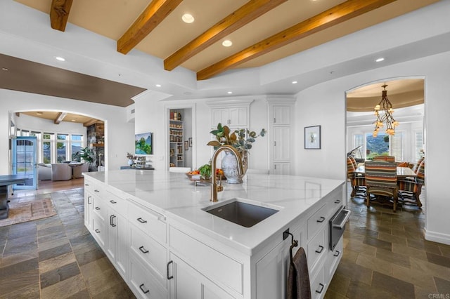 kitchen featuring beam ceiling, white cabinetry, sink, light stone counters, and a large island with sink