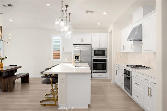 kitchen featuring appliances with stainless steel finishes, a kitchen island with sink, wall chimney range hood, decorative light fixtures, and white cabinetry