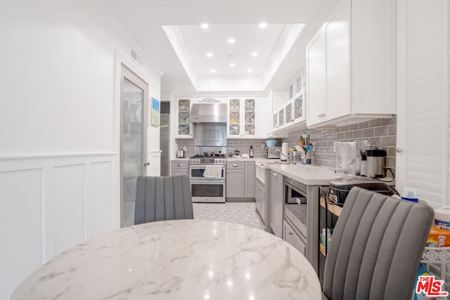 kitchen featuring double oven range, wall chimney exhaust hood, a raised ceiling, white cabinets, and light stone counters