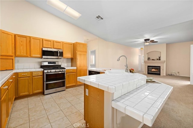 kitchen featuring tile countertops, a kitchen island with sink, sink, a fireplace, and appliances with stainless steel finishes