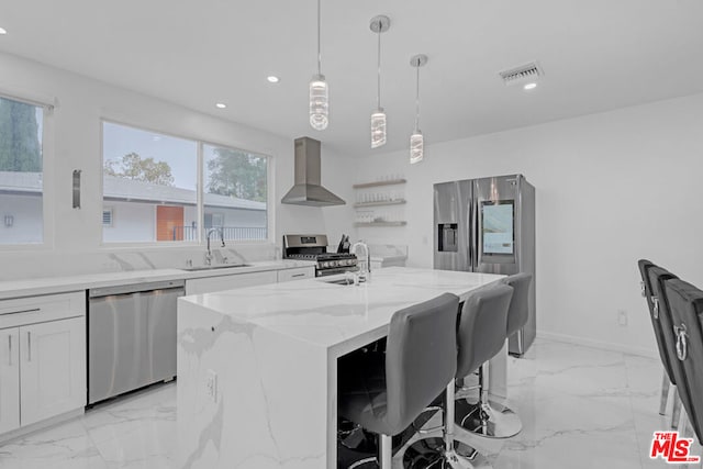 kitchen with wall chimney range hood, hanging light fixtures, a center island with sink, light stone countertops, and appliances with stainless steel finishes