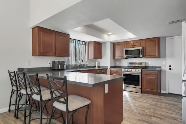 kitchen featuring appliances with stainless steel finishes, a tray ceiling, kitchen peninsula, a kitchen breakfast bar, and light hardwood / wood-style flooring