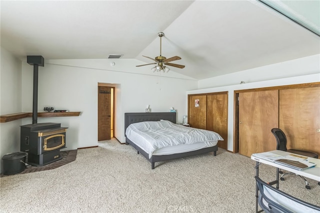 bedroom with a wood stove, vaulted ceiling, light colored carpet, two closets, and ceiling fan