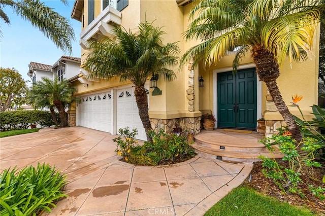 view of exterior entry featuring a tile roof, stucco siding, a garage, stone siding, and driveway