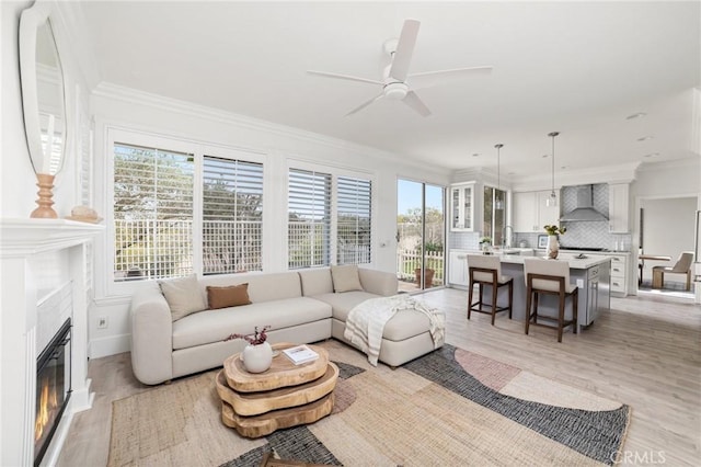 living room featuring a glass covered fireplace, crown molding, a ceiling fan, and light wood-style floors