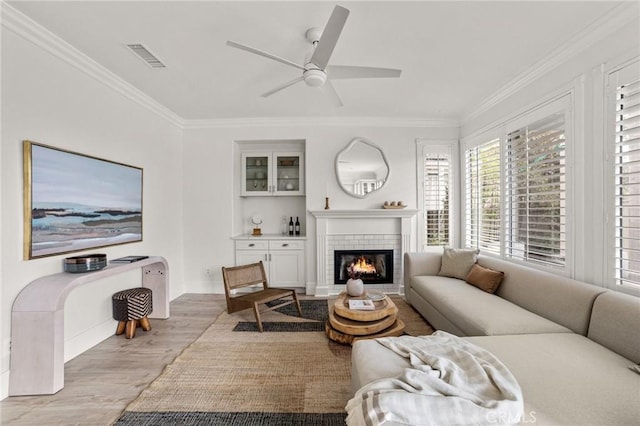 living room with visible vents, ornamental molding, a glass covered fireplace, light wood finished floors, and ceiling fan