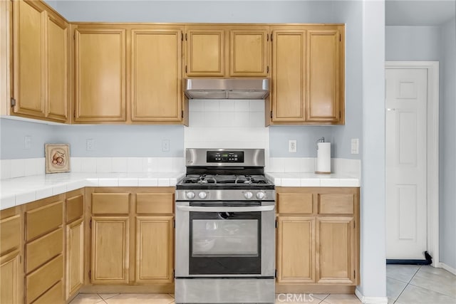 kitchen with tile counters, light brown cabinetry, stainless steel gas range oven, and light tile patterned flooring