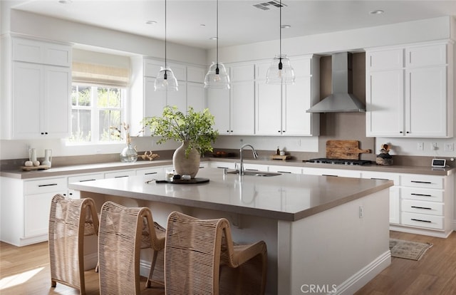 kitchen featuring wall chimney exhaust hood, an island with sink, sink, light wood-type flooring, and white cabinets