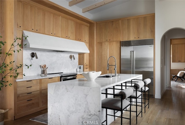 kitchen featuring beam ceiling, an island with sink, stainless steel appliances, sink, and light hardwood / wood-style floors