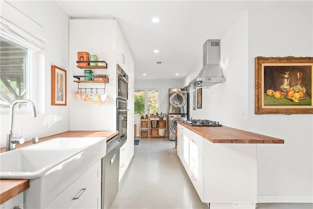 kitchen featuring butcher block counters, sink, wall chimney exhaust hood, stacked washer / drying machine, and white cabinets