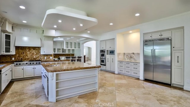 kitchen featuring built in appliances, an island with sink, tasteful backsplash, white cabinetry, and custom range hood