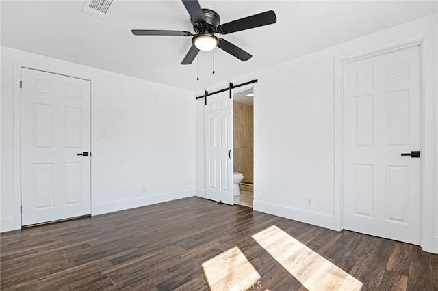 unfurnished bedroom featuring ensuite bath, a barn door, dark hardwood / wood-style flooring, and ceiling fan