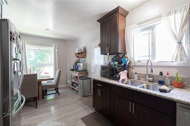 kitchen with sink, appliances with stainless steel finishes, dark brown cabinetry, light stone countertops, and light wood-type flooring