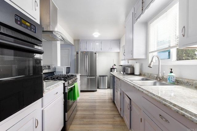 kitchen with appliances with stainless steel finishes, light wood-type flooring, sink, wall chimney range hood, and white cabinets